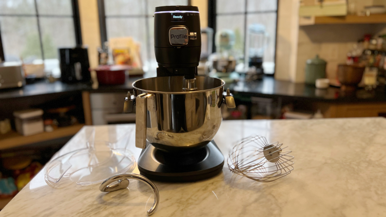 The GE Profile Smart Mixer sits on a marble table with the dough hook, whisk attachment and bowl shield on the table.