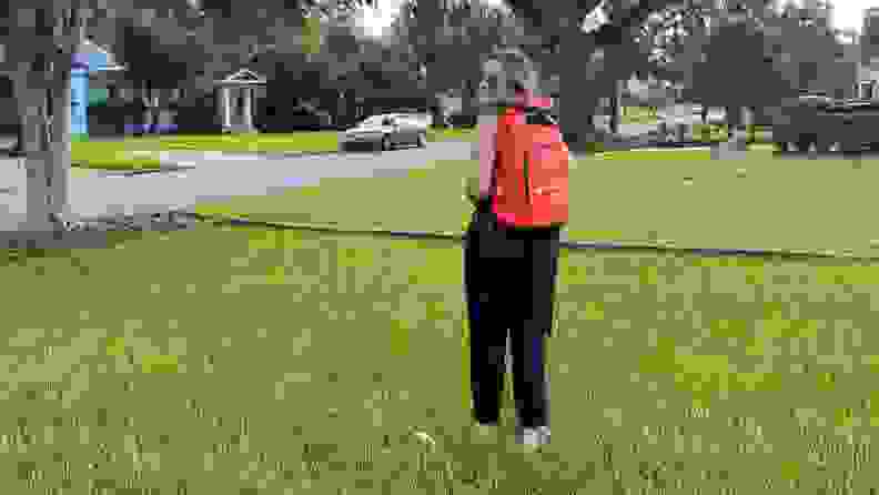 A person stands in the yard wearing an orange Judy backpack.