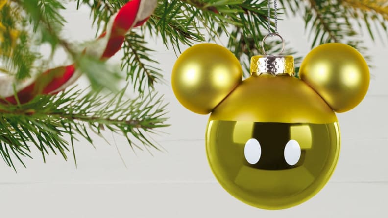 An image of a Disney Mickey Mouse holiday ornament in yellow dangling from a branch of a Christmas tree.