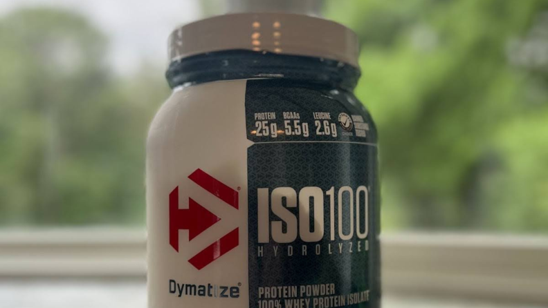 Close-up view of a bottle of Dymatize protein powder in front of a lush green background.