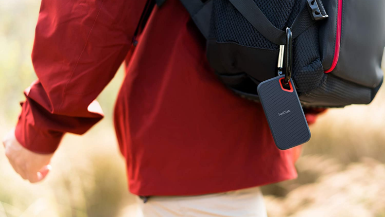 A person hikes with a SanDisk external hard drive clipped to their backpack.