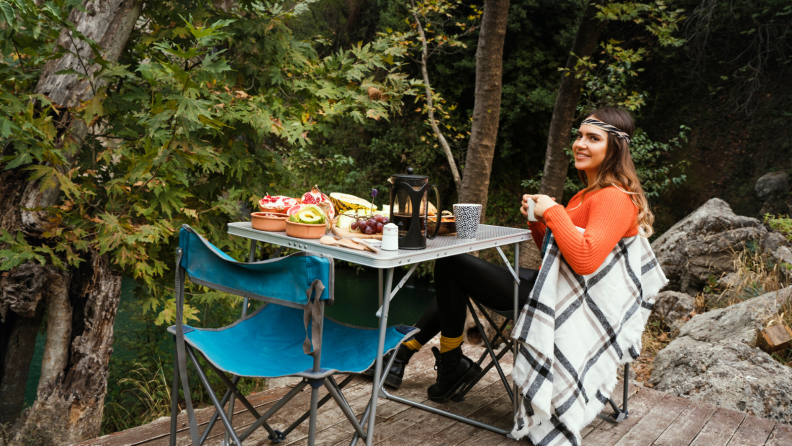 A woman is sitting in her foldable chair next to a foldable table in the woods. On the table, there are some food and a large pot of French press coffee. An empty folding chair is to the left of the table.