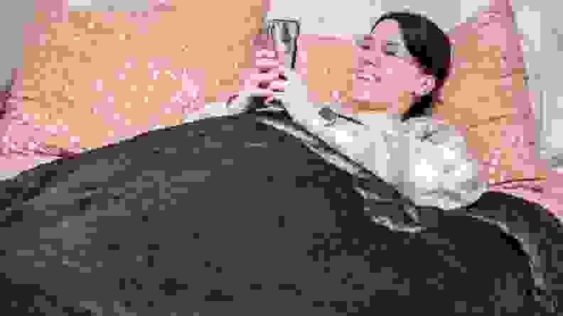 woman underneath navy blanket on bed using her phone