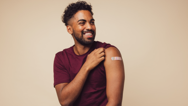 Man against brown background smiling after receiving vaccination, showing his arm with a bandage at point of injection