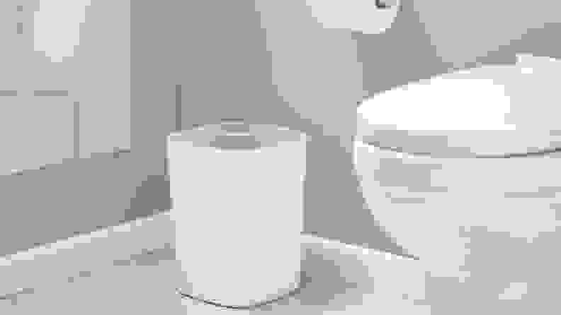 A all-gray bathroom contains a white toilet, a roll of toilet paper, and a trash can with an opening for trash and an opening for recycling.