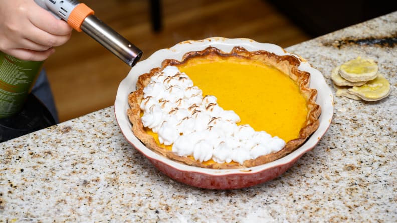 A hand using a butane torch to scorch meringue atop a pumpkin pie.