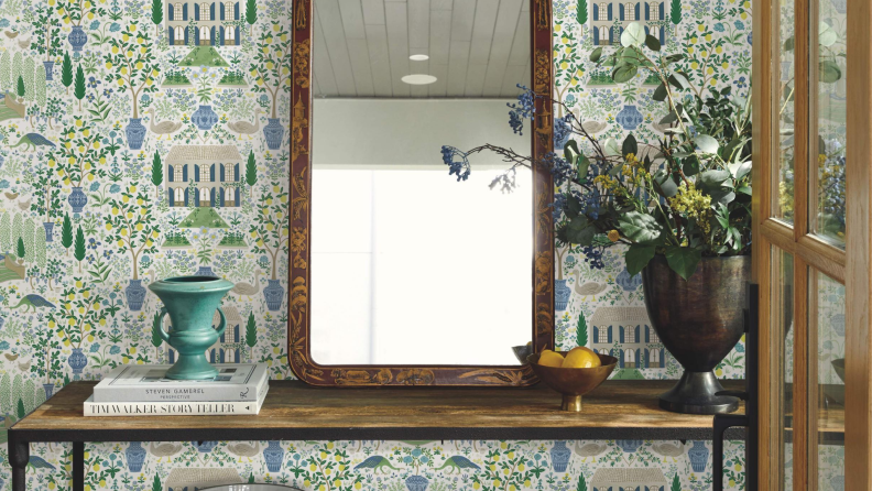 White, blue, yellow, and green wallpaper featuring a small vintage house next to fig and lemon trees. A wooden table with a mirror sits in front of the wallpaper