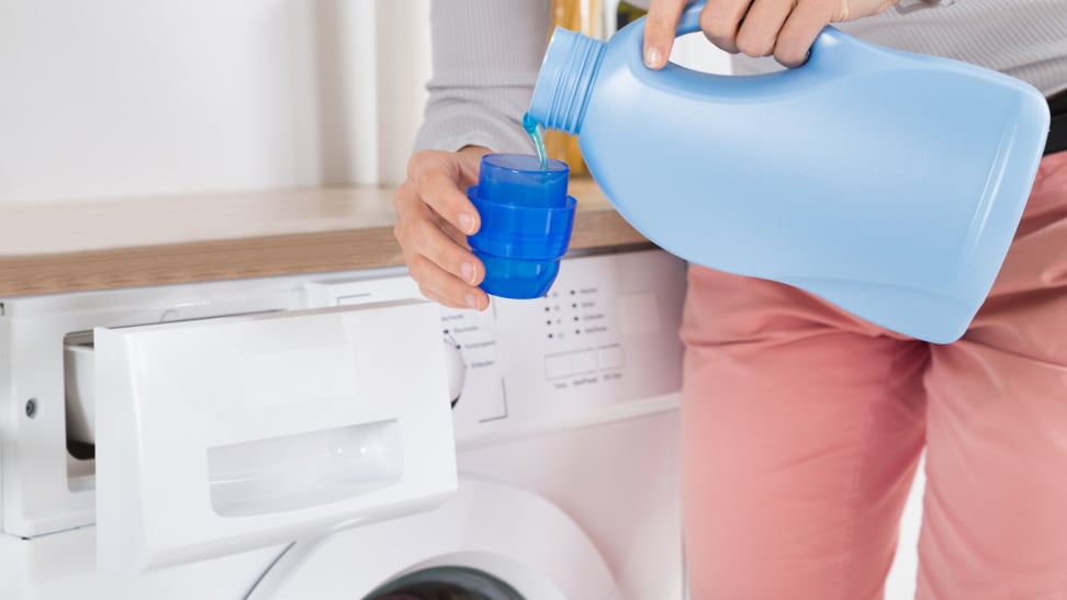 Person pouring liquid laundry detergent in cup in front of washing machine.