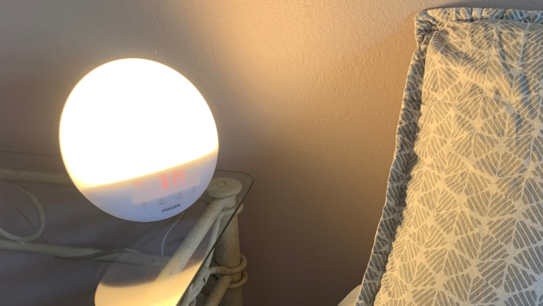 Image of the Philips SmartSleep sunrise alarm at full brightness in a room on a clear end table next a bed with a grey pillow