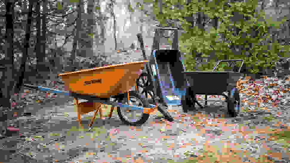 Three wheelbarrows—orange, black, and green—sit in a wooded area.