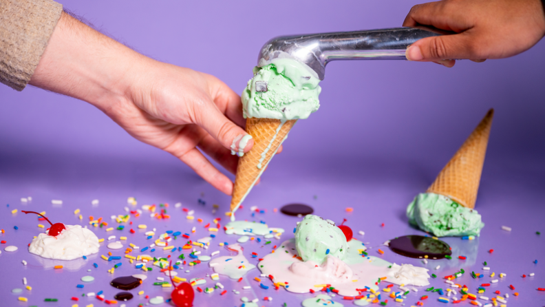 One hand holding a dripping ice cream cone, while another hand fills the cone with mint chocolate chip ice cream using the Zeroll.