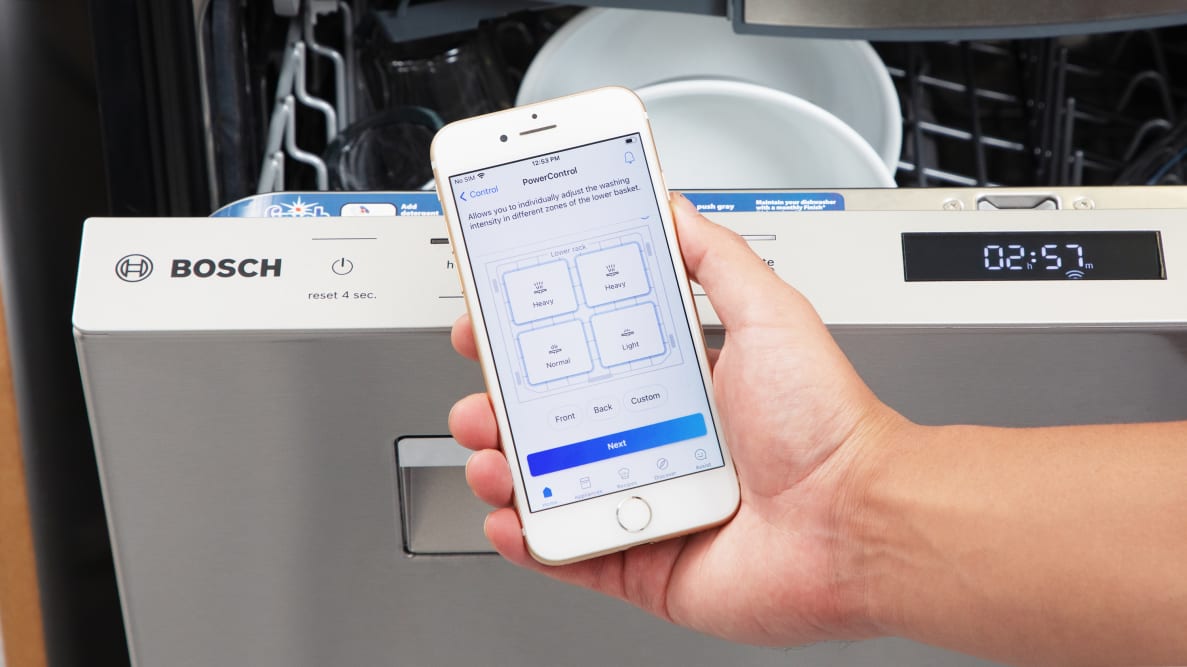 A person holds a phone showing the Home Connect app with four quadrants to control the Bosch 800 Series dishwasher, which is slightly open with dishes inside.