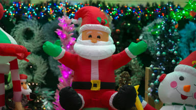A light-up inflatable Santa decoration surrounded by other Christmas iconography: trees, colorful Christmas lights, an inflatable snowman, white wreaths, and more.