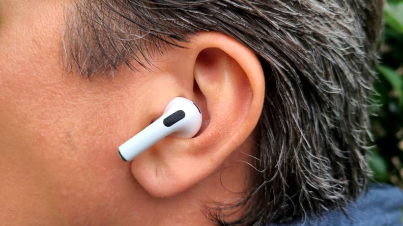 The all-white AirPod sits in the reviewer's ear with its shortened stem facing forward, set above a gret sweatshirt hood and below brown grey hair.