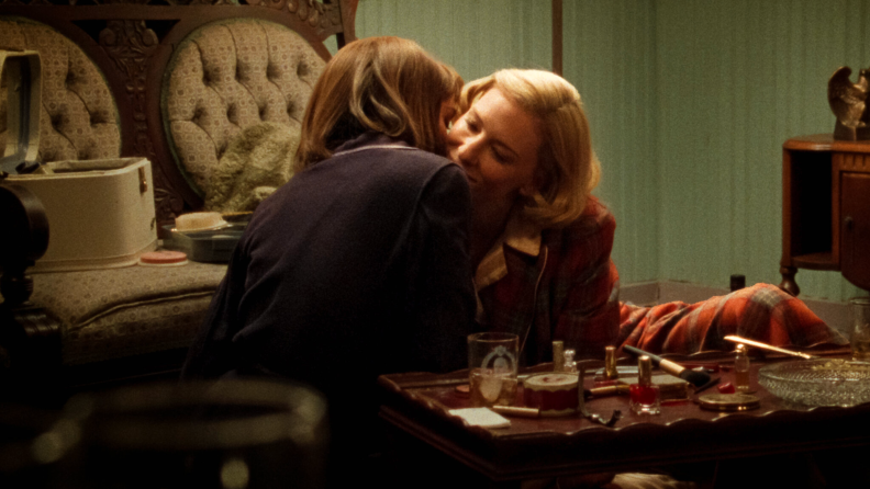 In Carol (2015), Cate Blanchett plays Carol Aird, leaning in to kiss another woman over drinks and a coffee table full of beauty products.