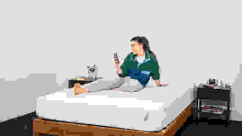 A woman sits on the Tuft & Needle Mattress on her phone