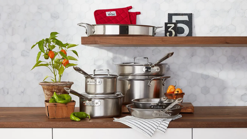 An AllClad cookware set on a wood counter next to a plant.