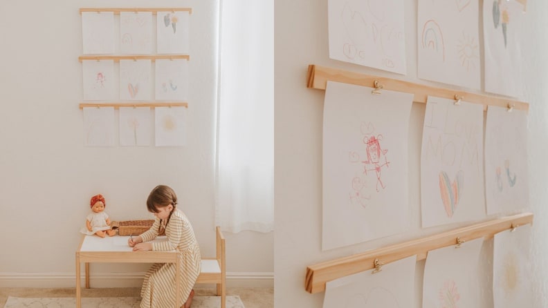 A small child sits drawing at a table. Above them: three horizontal wooden racks with metal clips, each displaying a piece of the child's art.