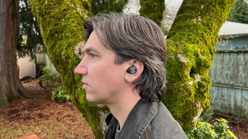 The gunsmoke colored wireless earbuds sit in a man's ear with a mossy tree in the background.