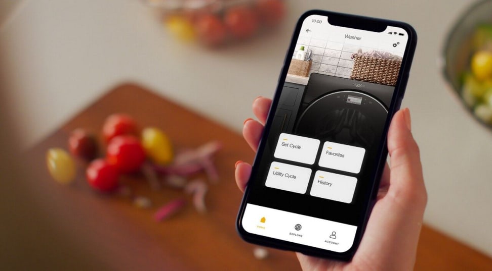 A hand holds a smart phone above a cooking board with some chopped vegetables on it. The phone's screen shows the Whirlpool app, and the user is checking on the status of their Whirlpool washing machine.