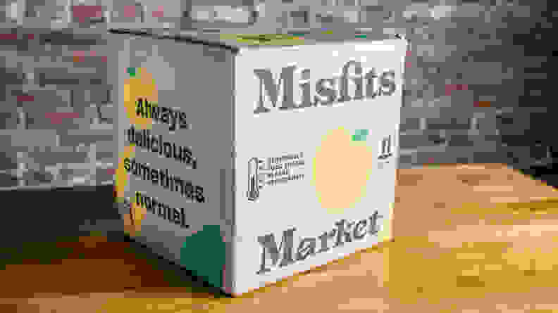 On a wooden tabletop, there's a Misfits Market box, potentially carrying produce items with imperfections.