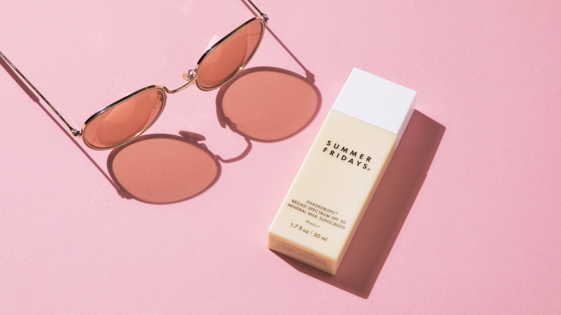A cream-colored, rectangular bottle of sunscreen lays on a pink backdrop with sunglasses next to it.