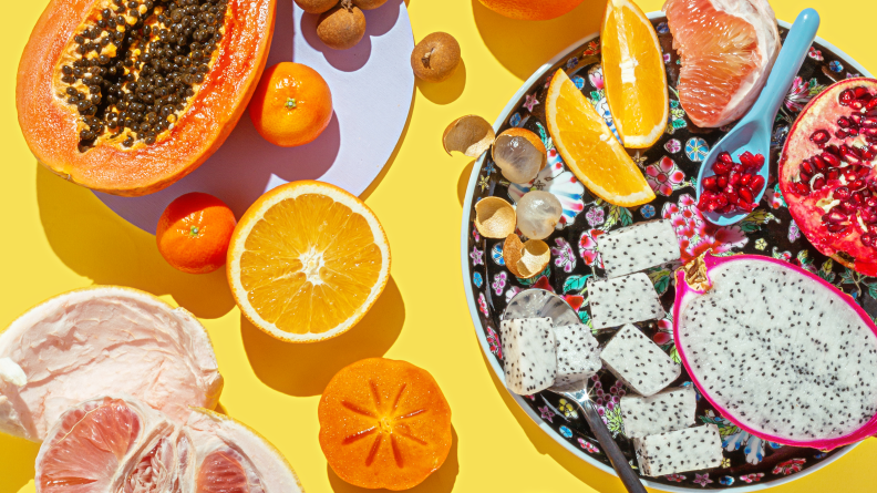 On a table, there's a large pink pomelo, a papaya, some oranges, and a dragon fruit.