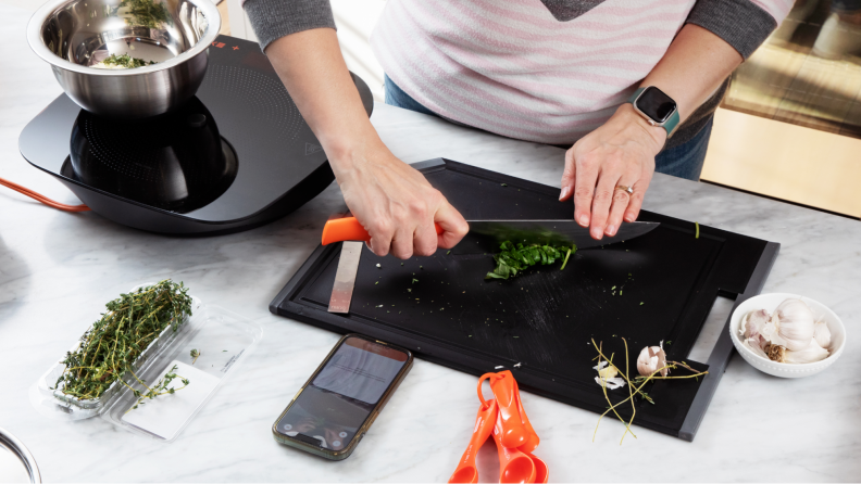 A person cutting some ingredients on a cutting board while looking at tutorial videos on a smartphone.