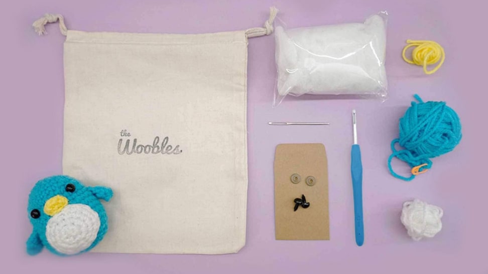 A crochet kit from the Woobles sits opened on a purple background, showcasing a stuffed penguin, crochet hook, yarn, and stuffing