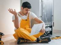A man sits on the ground near a fridge, near a drill and some manuals. He's reading the fridge's warranty and putting up one hand in confusion at what he's reading.