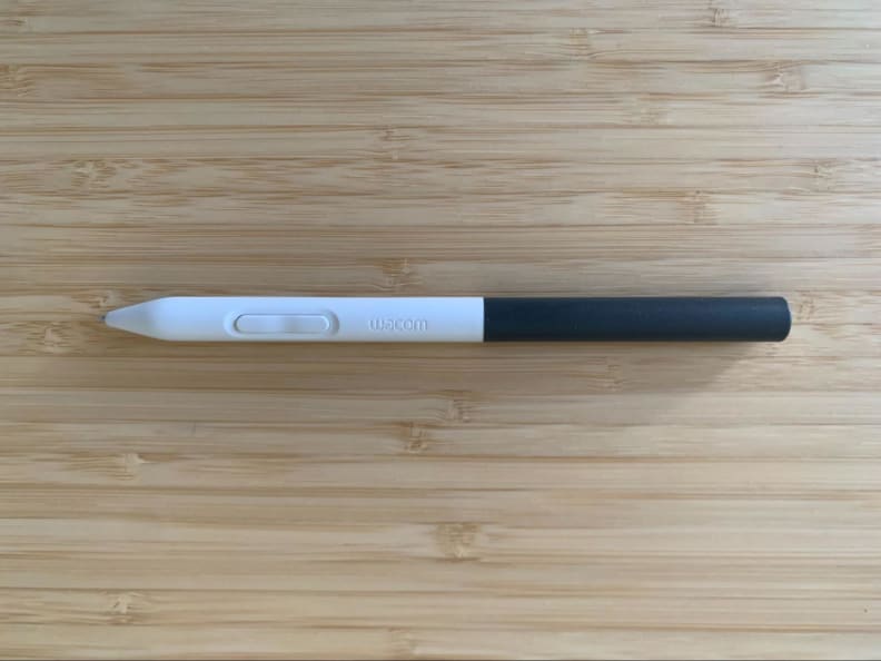 The white and gray Wacom digital drawing tablet pen appears on a bamboo desk, showing its button.