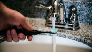 A man rinses the head of a black electric toothbrush under a bathroom faucet.