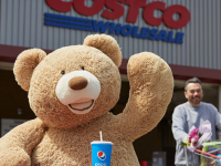 A large teddy bear is standing in front of a Costco while a shopper pushes a cart nearby. The bear has a pepsi cup.