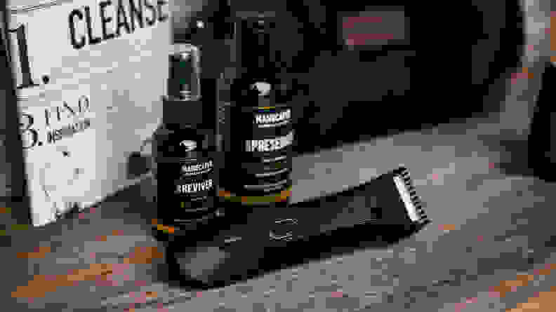 A beard trimmer laying on a table next to bottles of grooming products.