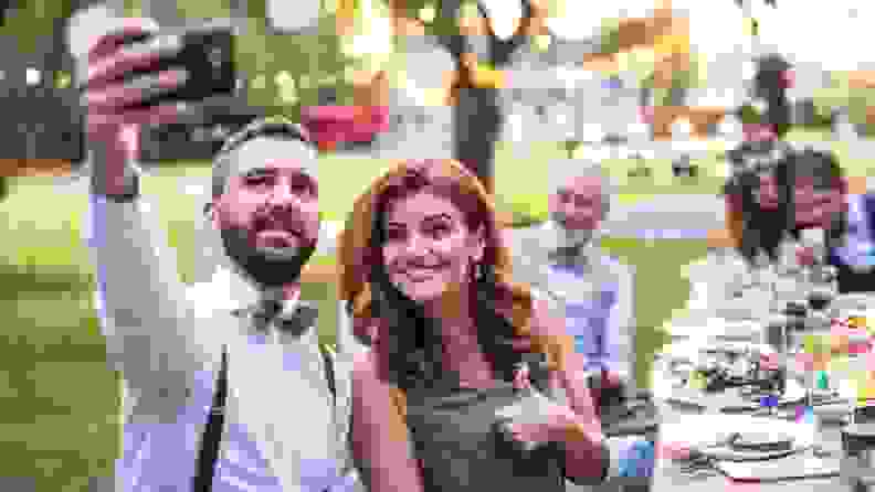 A wedding guest wearing suspenders and a bow tie takes a selfie with his date at the dinner table