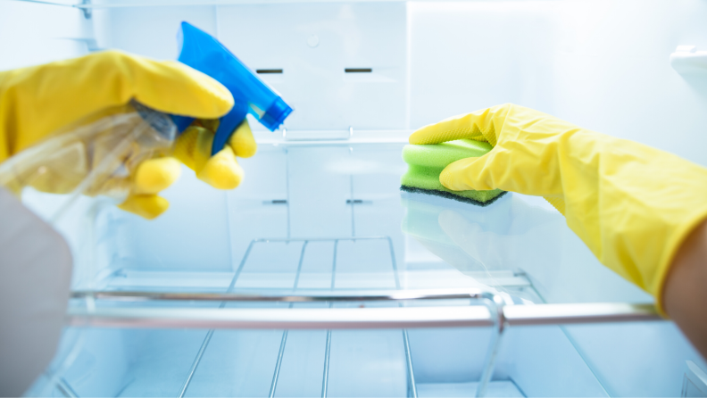 A point-of-view shot where the subject is wearing yellow rubber gloves and cleaning the inside of a fridge with a sponge.