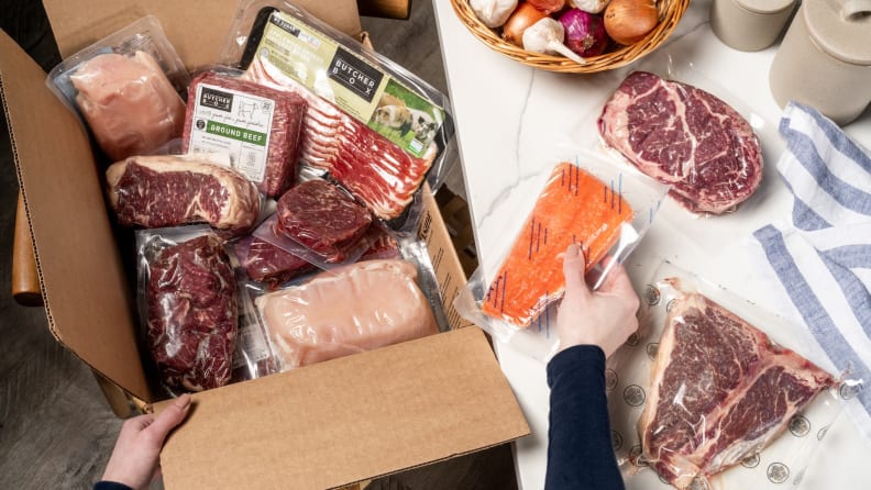ButcherBox meat delivery box filled with assorted vacuum-sealed cuts of beef, chicken, bacon, and salmon being unpacked on a kitchen counter.