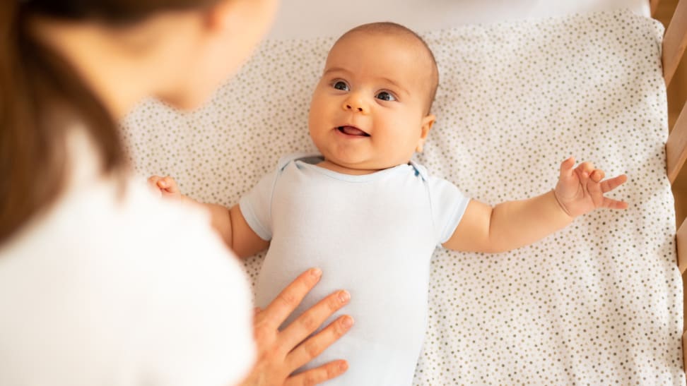 A baby smiles in their crib while their mom looks down on them.