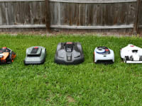 Five robot lawn mowers lined up together in the green grass with a fence behind