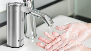 A pair of light-skinned hands are lathered with foamy soap under a running faucet. There is a Secura hand-soap dispenser in the foreground.