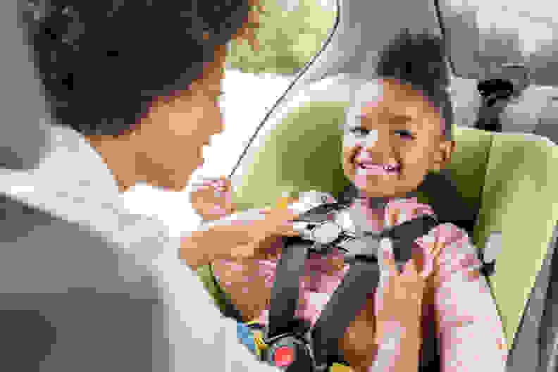 A young smiling girl getting her car seat straps adjusted by her mother in a car.