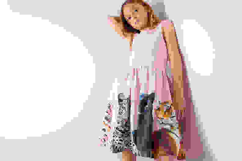 A girl wearing a pink dress printed with cats leans against a wall.