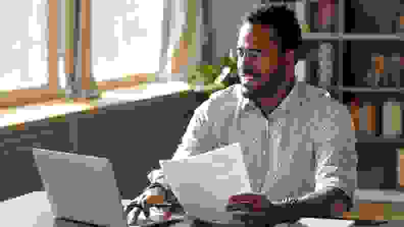 A man wearing glasses sits at his laptop holding a financial document