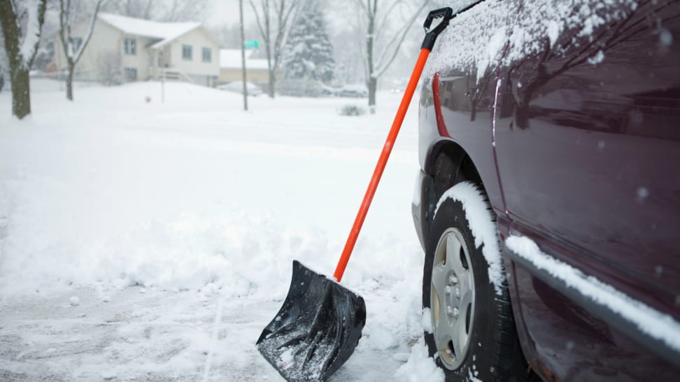 Snow shovel propped up on a dark car covered in snow, surrounded by snow on the ground