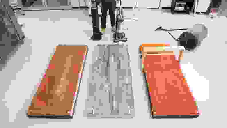 A man in a testing lab stands with a vacuum in front of three different vacuum testing surfaces including medium-pile and high-pile carpet, hard flooring