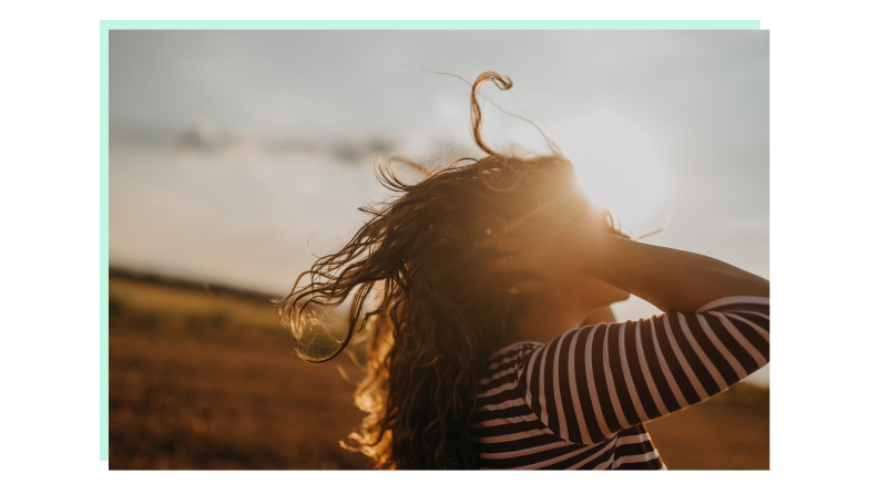 The silhouette of a person running their hands through their long curly hair with the sun setting in the background.