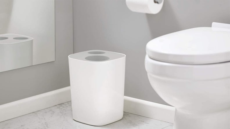 A all-gray bathroom contains a white toilet, a roll of toilet paper, and a trash can with an opening for trash and an opening for recycling.