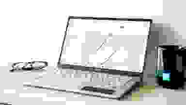 An open and powered on silver laptop sitting on a white surface with a black mug on the right and a pair of glasses on the left