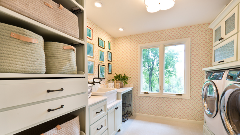 This photograph shows how wire, woven, or rattan baskets can help your laundry room look chic and serve a purpose. Decorative pictures are hung on the wall opposite a washer and dryer; the sun is shining outside the window.