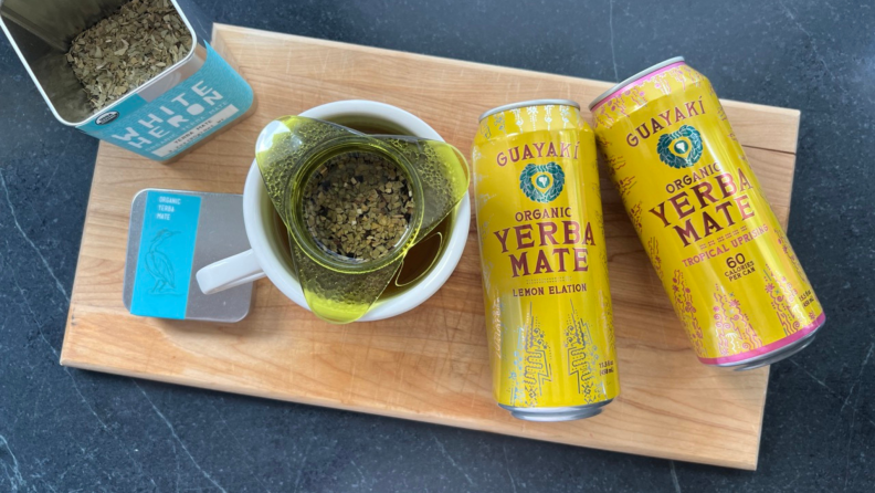 A can of yerba mate leaves sites next to a mug of steeping yerba mate, and 2 cans of yerba mate.